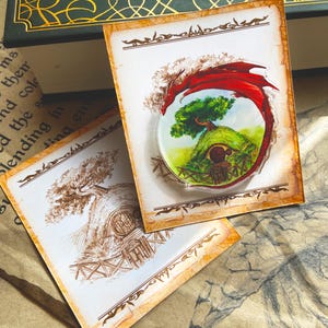Può includere: Una spilla rotonda in acrilico trasparente con una vivace illustrazione di una casa hobbit su una collina verde, incorniciata da un drago rosso. Sono visibili anche due biglietti di carta invecchiata con disegni seppia di un albero e una casa hobbit.