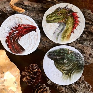 K&ouml;nnte beinhalten: Drei runde Aufkleber mit detaillierten Illustrationen von Drachen. Der erste Aufkleber zeigt einen roten Drachen vor wei&szlig;em Hintergrund. Der zweite Aufkleber zeigt einen gr&uuml;nen Drachen vor wei&szlig;em Hintergrund. Der dritte Aufkleber zeigt einen gr&uuml;n-braunen Drachen vor wei&szlig;em Hintergrund.