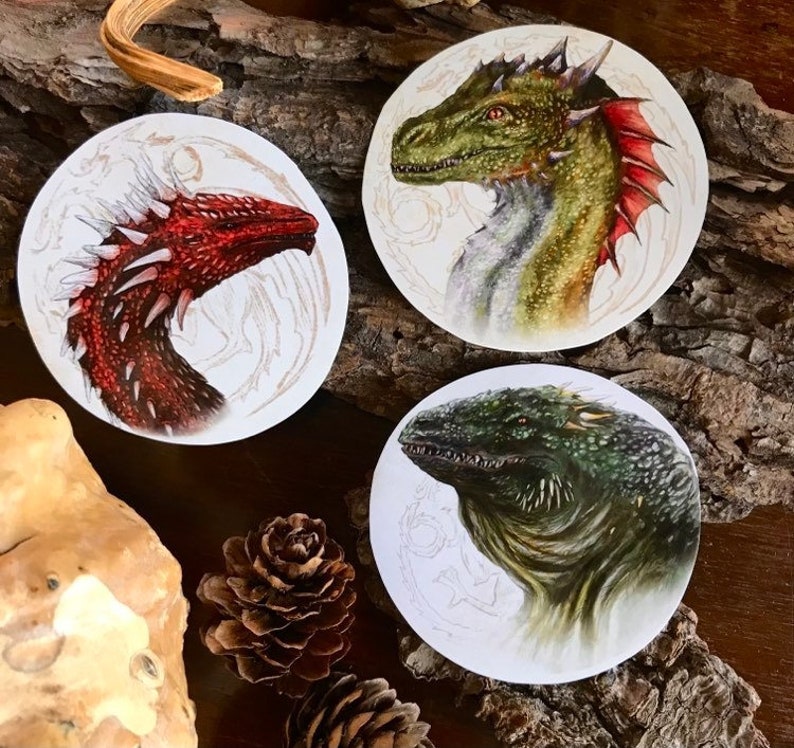 K&ouml;nnte beinhalten: Drei runde Aufkleber mit detaillierten Illustrationen von Drachen. Der erste Aufkleber zeigt einen roten Drachen vor wei&szlig;em Hintergrund. Der zweite Aufkleber zeigt einen gr&uuml;nen Drachen vor wei&szlig;em Hintergrund. Der dritte Aufkleber zeigt einen gr&uuml;n-braunen Drachen vor wei&szlig;em Hintergrund.