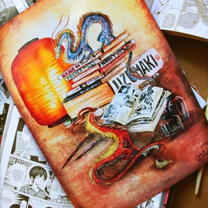 Può includere: Illustrazione di un drago rosso e giallo e di un drago blu e bianco, con una lanterna arancione e una pila di libri. I draghi sono vicino a un fumetto aperto. L'immagine include il testo "IZUMAKI".