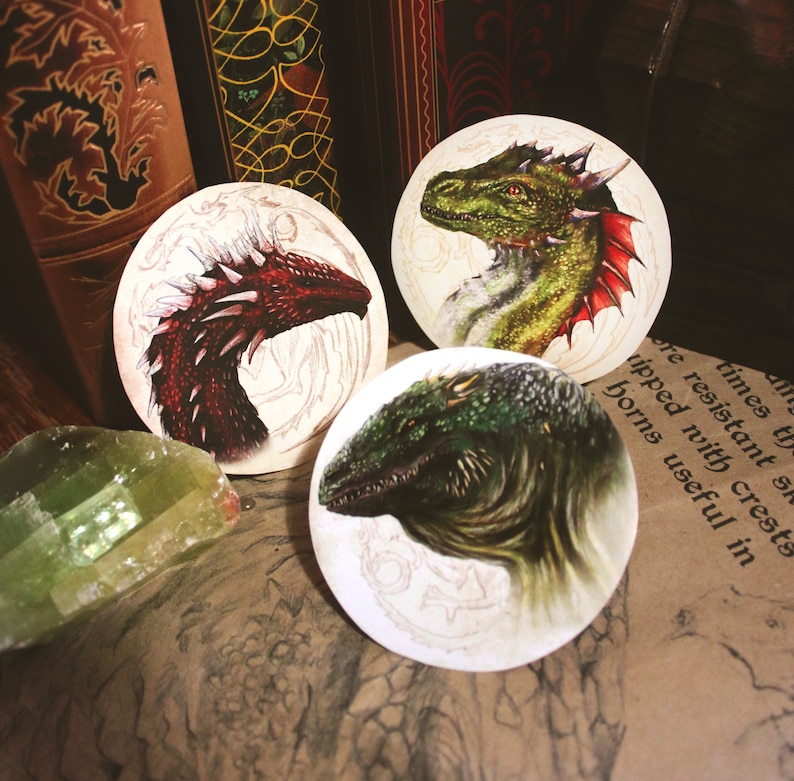 K&ouml;nnte beinhalten: Drei runde Untersetzer mit Illustrationen von Drachen. Der erste Untersetzer zeigt einen roten Drachen vor wei&szlig;em Hintergrund. Der zweite Untersetzer zeigt einen gr&uuml;nen Drachen vor wei&szlig;em Hintergrund. Der dritte Untersetzer zeigt einen gr&uuml;n-braunen Drachen vor wei&szlig;em Hintergrund.