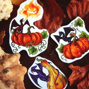May include: Set of three stickers featuring pumpkins and dragons. One sticker shows a dragon on a pumpkin with a fiery face above. Another shows a black dragon on a textured pumpkin. The third shows a purple dragon on a long, orange pumpkin.