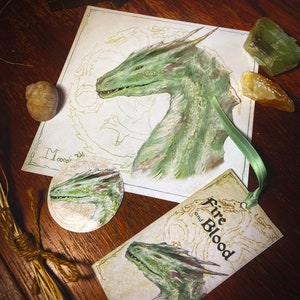 May include: An artistic composition featuring a green dragon illustration on a print, a bookmark, and a small round object. The bookmark reads "Fire and Blood." The dragon art is detailed with green and brown hues. The items are arranged on a wooden surface.
