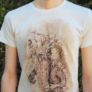 Op de afbeelding: Crèmekleurig T-shirt met een gedetailleerde bruine illustratie van een boomachtige figuur met een lange baard en ingewikkelde takken. Het kunstwerk heeft een aquareleffect. Het shirt heeft een ronde hals en korte mouwen.