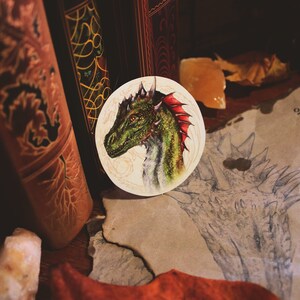 K&ouml;nnte beinhalten: Eine Nahaufnahme einer gr&uuml;nen und roten Drachenkopf-Illustration auf einem wei&szlig;en runden Aufkleber. Der Aufkleber befindet sich auf einer braunen Oberfl&auml;che mit einer Zeichnung eines Drachen auf einem Blatt Papier.
