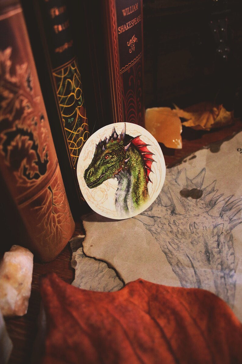 K&ouml;nnte beinhalten: Eine Nahaufnahme einer gr&uuml;nen und roten Drachenkopf-Illustration auf einem wei&szlig;en runden Aufkleber. Der Aufkleber befindet sich auf einer braunen Oberfl&auml;che mit einer Zeichnung eines Drachen auf einem Blatt Papier.