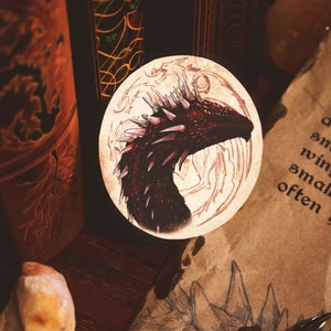 K&ouml;nnte beinhalten: Ein runder Aufkleber mit einer Illustration eines roten Drachenkopfes auf beigefarbenem Hintergrund. Der Drache hat scharfe Z&auml;hne und einen stacheligen Kamm. Der Aufkleber ist umgeben von einer braunen Holzoberfl&auml;che und einem St&uuml;ck Pergamentpapier.