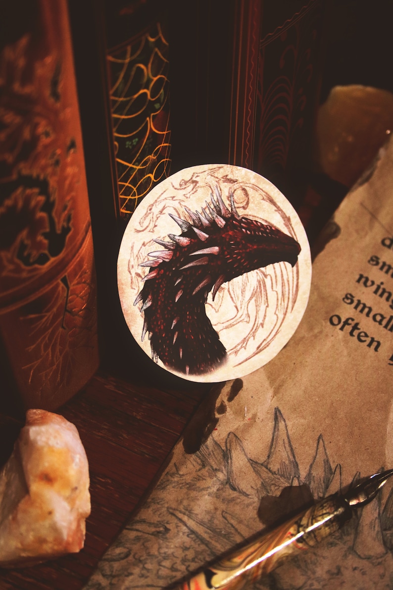 K&ouml;nnte beinhalten: Ein runder Aufkleber mit einer Illustration eines roten Drachenkopfes auf beigefarbenem Hintergrund. Der Drache hat scharfe Z&auml;hne und einen stacheligen Kamm. Der Aufkleber ist umgeben von einer braunen Holzoberfl&auml;che und einem St&uuml;ck Pergamentpapier.