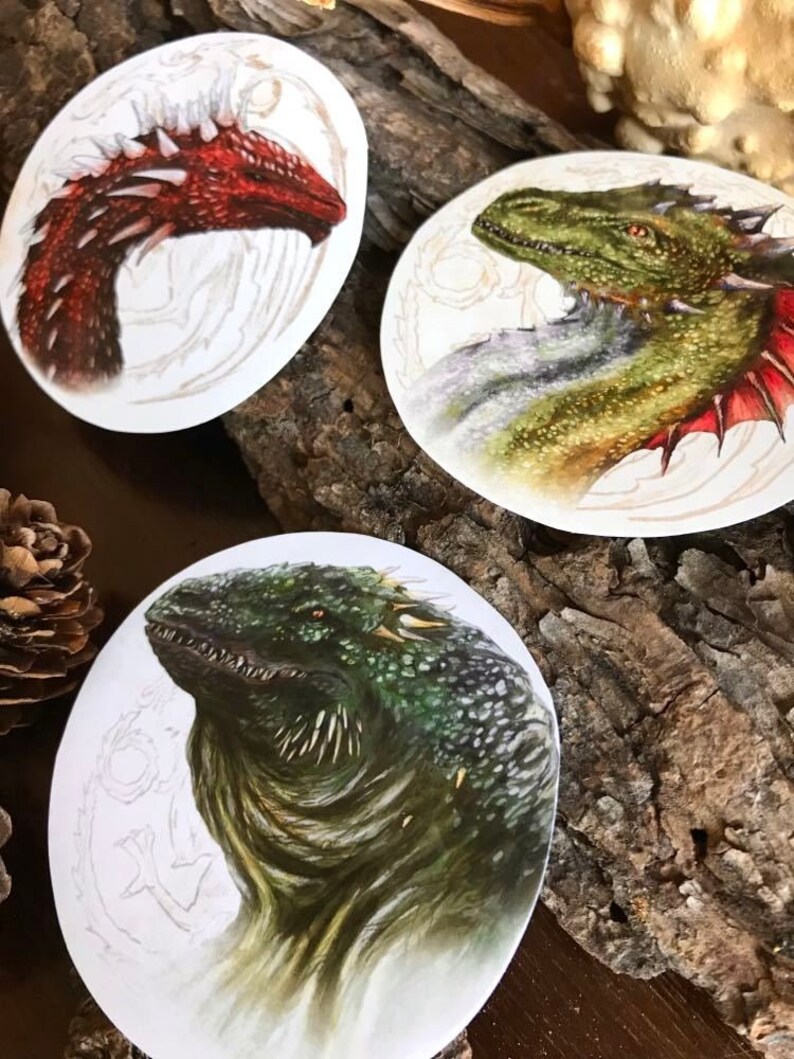 K&ouml;nnte beinhalten: Drei runde Aufkleber mit detaillierten Illustrationen von Drachen. Der erste Aufkleber zeigt einen roten Drachen mit wei&szlig;en Stacheln auf R&uuml;cken und Kopf. Der zweite Aufkleber zeigt einen gr&uuml;nen Drachen mit wei&szlig;en und roten Akzenten. Der dritte Aufkleber zeigt einen gr&uuml;nen Drachen mit einem reptilienartigen Aussehen.