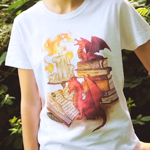 May include: White t-shirt with a fantasy-themed design. The illustration depicts two red dragons, books, and candles. One dragon is on a stack of books, another near an open book. The artwork is in a watercolour style.