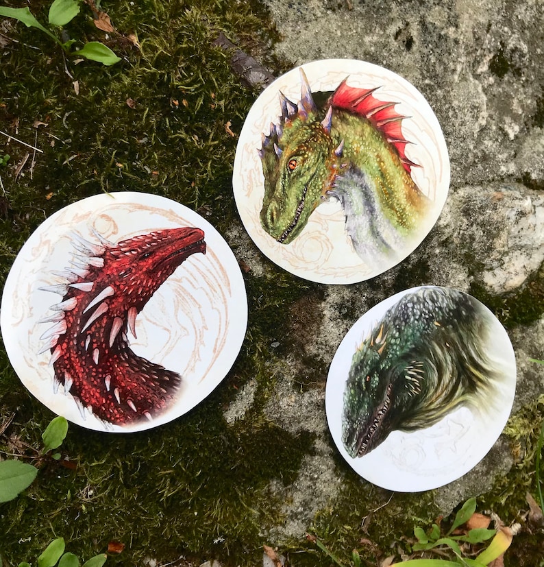 K&ouml;nnte beinhalten: Drei runde Aufkleber mit bunten Illustrationen von Drachen. Der erste Aufkleber zeigt einen gr&uuml;nen Drachen mit roten Akzenten, der zweite Aufkleber zeigt einen roten Drachen mit wei&szlig;en Akzenten und der dritte Aufkleber zeigt einen gr&uuml;nen und schwarzen Drachen.