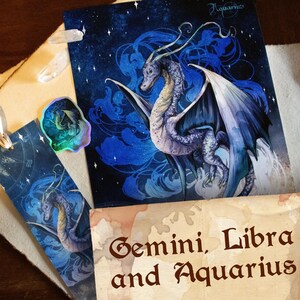 Può includere: Un'illustrazione di un drago blu e viola su uno sfondo di cielo stellato. L'opera è presente su una stampa, un segnalibro e un adesivo olografico. La stampa ha la parola "Aquarius". Il testo "Gemini, Libra and Aquarius" è in basso.