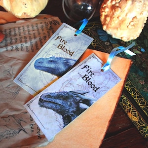 May include: Two rectangular bookmarks with the text "Fire and Blood" and a blue dragon illustration. Each bookmark has a blue ribbon attached. The background includes parchment and decorative elements.