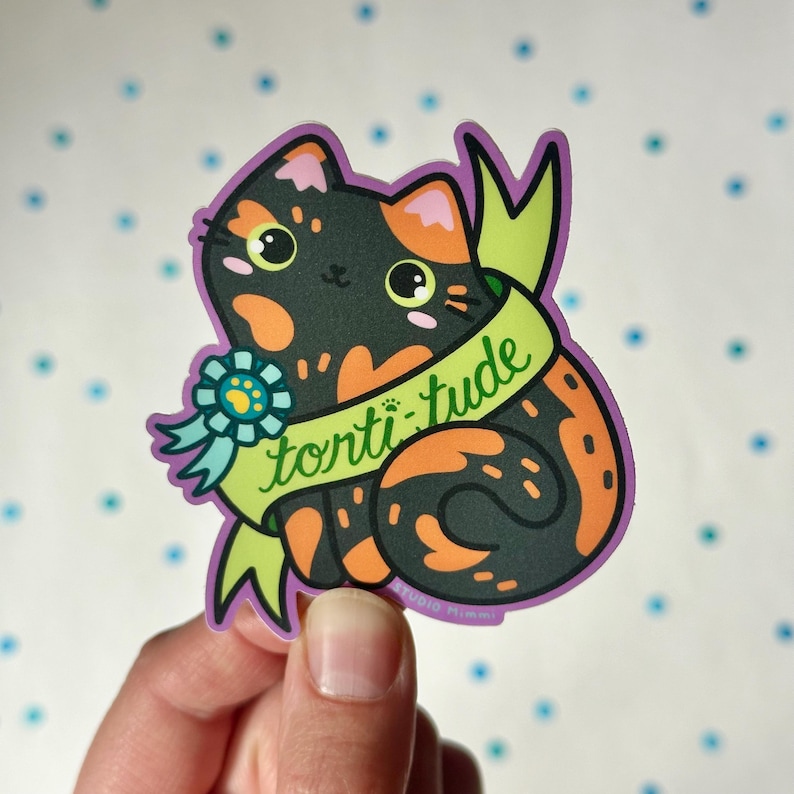 May include: A sticker featuring a cartoon calico cat with orange, black, and white markings. The cat is wearing a green ribbon with the text "tortitude" written on it. The sticker has a purple border.