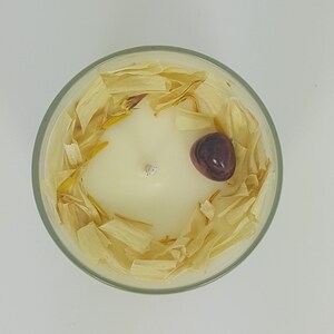May include: A top-down view of a scented candle in a clear glass container. The candle is ivory-colored with a single wick. It is decorated with dried, light-yellow petals and a dark-purple stone. The candle sits on a white surface.