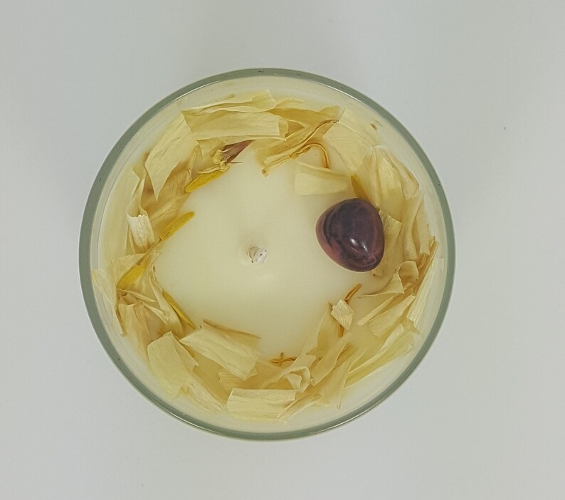 May include: A top-down view of a scented candle in a clear glass container. The candle is ivory-colored with a single wick. It is decorated with dried, light-yellow petals and a dark-purple stone. The candle sits on a white surface.