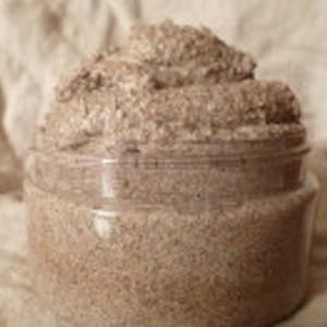 May include: A close-up of a brown sugar scrub in a clear glass jar. The scrub is a light brown color and has a grainy texture.