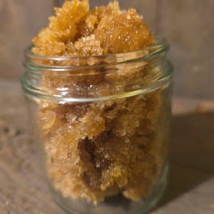 May include: A small, clear glass jar filled with a golden-brown, granular substance. The jar is sitting on a wooden surface. The contents appear to be a body scrub or similar cosmetic product.
