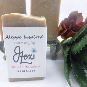 Op de afbeelding: Twee natuurlijke, handgemaakte zeepjes ge&iuml;nspireerd door Aleppo-zeep. De zeep is lichtbruin van kleur en heeft een etiket met de tekst "Aleppo Inspired Face + body by Lexi Natural + Handmade net wt 4.15 oz".