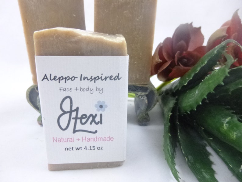 Op de afbeelding: Twee natuurlijke, handgemaakte zeepjes ge&iuml;nspireerd door Aleppo-zeep. De zeep is lichtbruin van kleur en heeft een etiket met de tekst "Aleppo Inspired Face + body by Lexi Natural + Handmade net wt 4.15 oz".