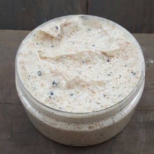 May include: A close-up of a white body scrub with black seeds in a clear jar. The scrub is thick and creamy, and the seeds are evenly distributed throughout.