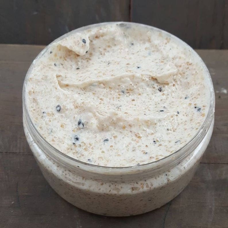 May include: A close-up of a white body scrub with black seeds in a clear jar. The scrub is thick and creamy, and the seeds are evenly distributed throughout.