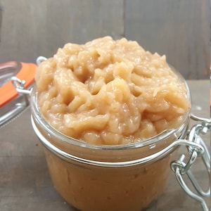Pode incluir: Um frasco de vidro transparente cheio de um alimento castanho claro e granulado, possivelmente puré de maçã ou uma conserva semelhante. O frasco tem um fecho de metal e uma vedação de borracha laranja. O fundo é uma superfície de madeira.