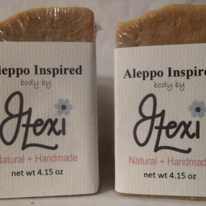 Op de afbeelding: Twee repen Aleppo Inspired zeep van Hexi. Elke reep is verpakt in wit papier met de merknaam en productdetails in zwart gedrukt. De zeep is lichtbruin van kleur en heeft een natuurlijke, handgemaakte uitstraling.