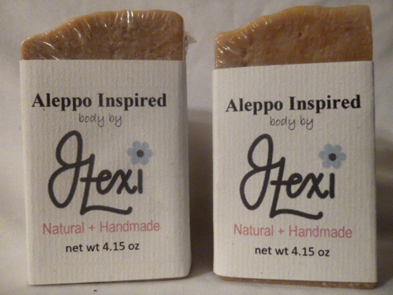 Op de afbeelding: Twee repen Aleppo Inspired zeep van Hexi. Elke reep is verpakt in wit papier met de merknaam en productdetails in zwart gedrukt. De zeep is lichtbruin van kleur en heeft een natuurlijke, handgemaakte uitstraling.