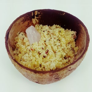 May include: A close-up of a coconut shell bowl filled with a yellow sugar scrub. A small pink crystal is nestled in the scrub.