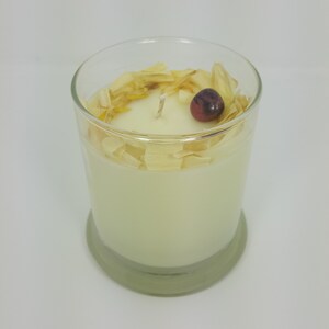 May include: A scented candle in a clear glass container. The candle is ivory-colored and topped with dried petals and a small, dark red berry. The glass has a rounded base and a thick rim.