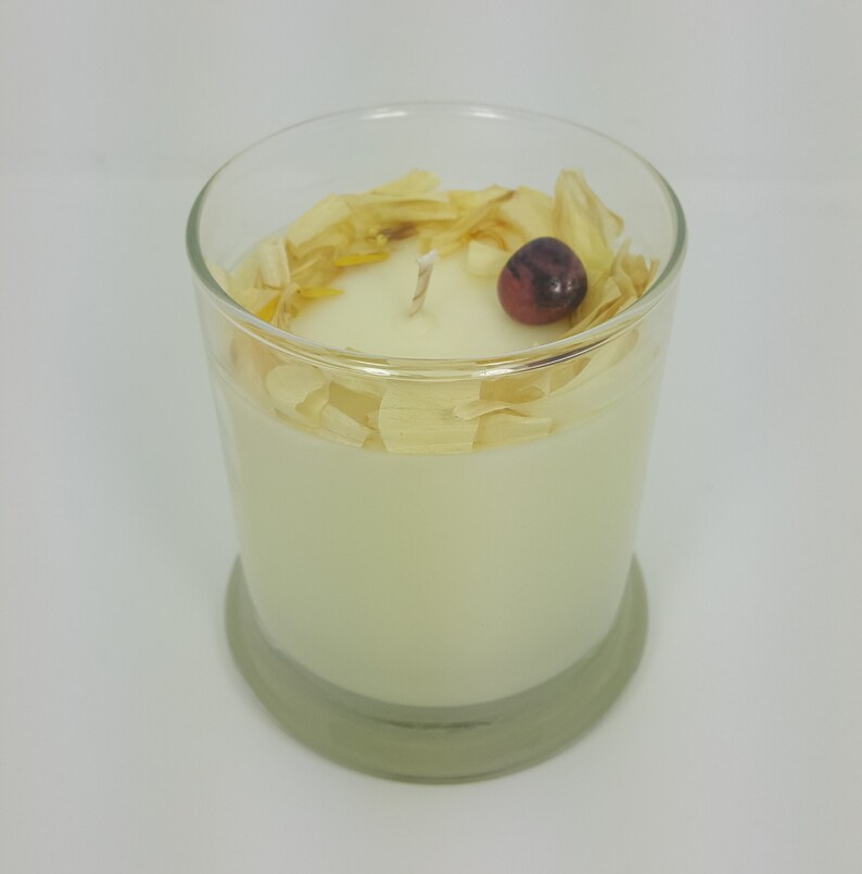 May include: A scented candle in a clear glass container. The candle is ivory-colored and topped with dried petals and a small, dark red berry. The glass has a rounded base and a thick rim.