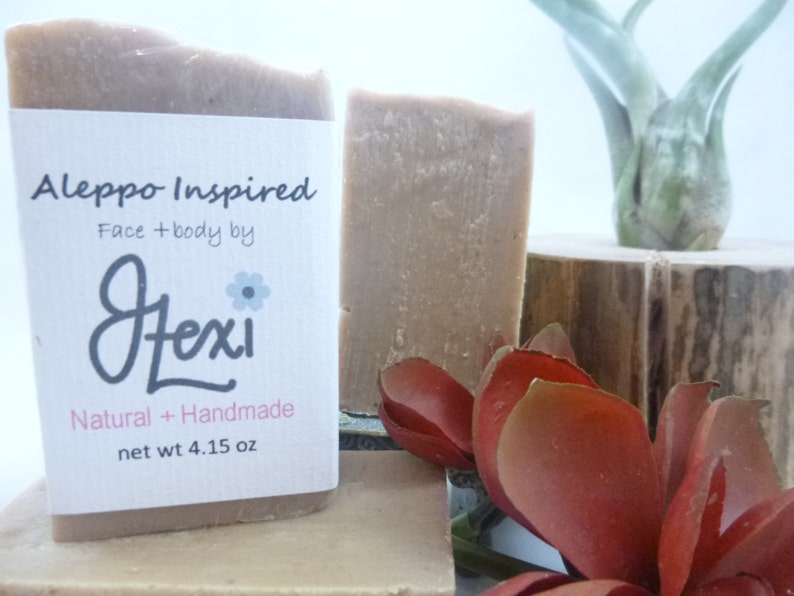 Op de afbeelding: Een Aleppo-ge&iuml;nspireerde, natuurlijke, handgemaakte zeep van Jexi, met een wit etiket waarop staat "Aleppo Inspired Face + body by Jexi Natural + Handmade net wt 4.15 oz".