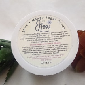 May include: White jar with a pink and black label that reads "Shea + Mango Sugar Scrub by Hexi". The label includes instructions for use and a list of ingredients. The jar is sitting on a green and brown surface.