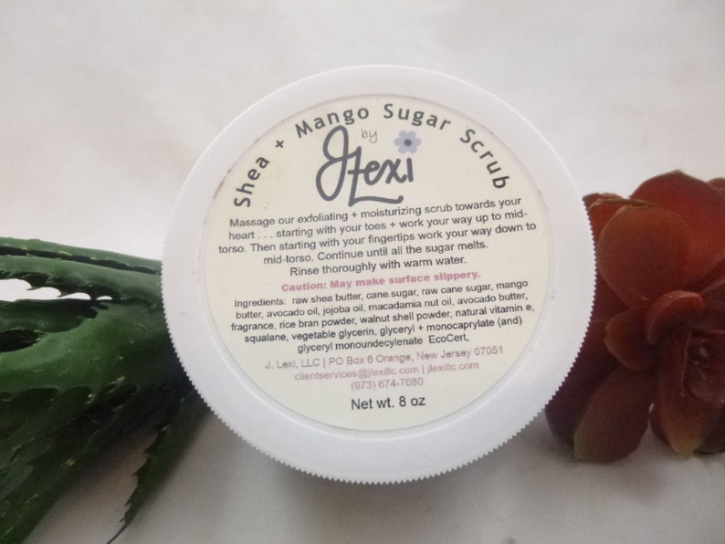 May include: White jar with a pink and black label that reads "Shea + Mango Sugar Scrub by Hexi". The label includes instructions for use and a list of ingredients. The jar is sitting on a green and brown surface.