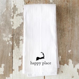 May include: White kitchen towel with a black graphic of a coastline and the words "happy place" printed below. The towel is folded and has a slightly wrinkled texture. The background is a weathered wooden surface.