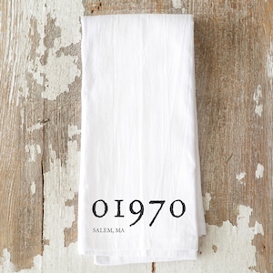 May include: White kitchen towel with the black text "01970" and "SALEM, MA" printed on it. The towel is folded and lies on a rustic wooden surface.