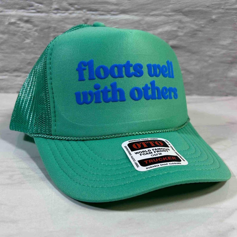May include: A bright green trucker hat featuring a foam front panel with the blue text "floats well with others" printed in a bold font. The cap includes a mesh back, a curved brim, and a white sticker on the brim with black text.
