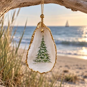 May include: A decorative oyster shell ornament featuring a hand-painted green Christmas tree on the interior. The shell edges are trimmed in gold, and it hangs from a gold-toned cord with a small gold star charm attached at the top of the shell.