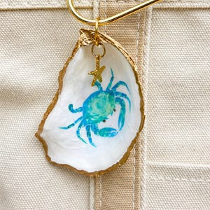 May include: A decorative oyster shell ornament featuring a hand-painted blue and green crab on the interior. The shell edges are trimmed in gold paint, and a small gold-toned metal starfish charm hangs from a gold jump ring at the top.