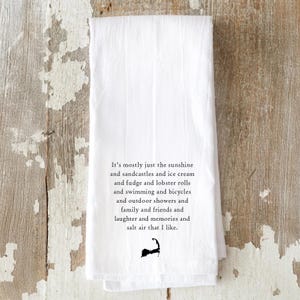 May include: White kitchen towel with black text that reads "It's mostly just the sunshine and sandcastles and ice cream and fudge and lobster rolls and swimming and bicycles and outdoor showers and family and friends and laughter and memories and salt air that I like." A black silhouette of a land mass is at the bottom.