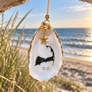 May include: A decorative oyster shell ornament with a gold-painted edge, hanging from a gold cord. The interior features a black silhouette map of Cape Cod, the letters C.C., and a small gold starfish charm suspended from the top jump ring.