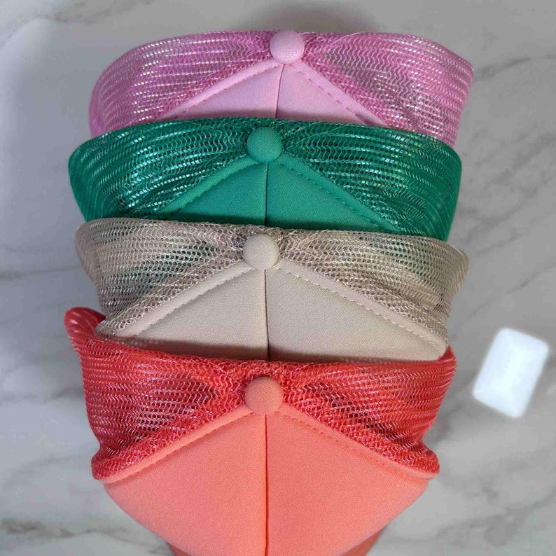 May include: A stack of four trucker hats featuring a solid foam front panel and a breathable mesh back panel. The hats are colored in pink, green, beige, and coral, each topped with a matching fabric-covered button at the center of the crown.