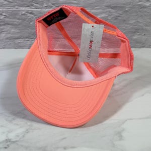 May include: A bright neon coral trucker hat with a mesh back and a solid front panel. The interior features a black tag with the text OTTO and a white paper hang tag with the text lauralewdesign printed in red and black lettering.