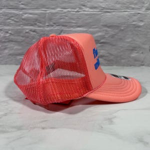 May include: A coral-colored trucker hat featuring a mesh back panel and a solid front panel with the blue text "float with" printed on the side. The cap includes a curved brim and a structured foam front, showcasing a vibrant monochromatic design.