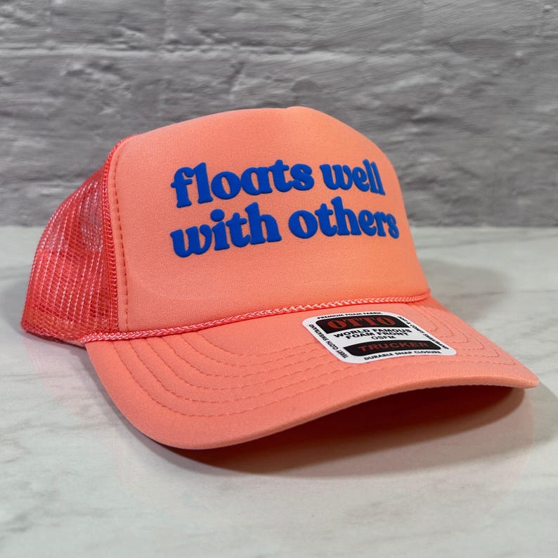 May include: A neon orange trucker hat featuring a mesh back and a foam front panel. The text "floats well with others" is printed in bold blue letters on the front. A small rectangular product label is attached to the brim of the hat.