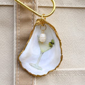 May include: A decorative oyster shell ornament featuring a painted martini glass with two green olives. The shell has a gold-painted rim and is suspended from a gold-toned metal hook. A small, white, oval-shaped pearl charm hangs from a gold ring.
