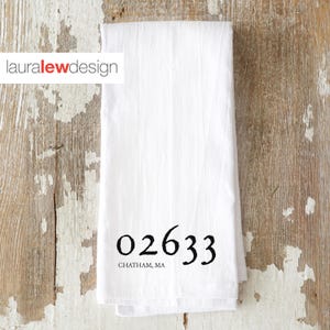 May include: White folded kitchen towel with the text "02633 CHATHAM, MA" in black. The towel has the "lauralewdesign" logo in the upper left corner. The towel is on a distressed wooden surface.