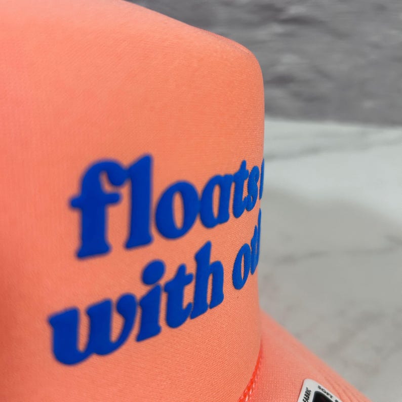May include: A bright coral-colored trucker hat featuring the blue text "floats with o" printed on the front panel. The hat has a structured foam front and a visible seam along the side, showcasing a bold color contrast between the fabric and text.