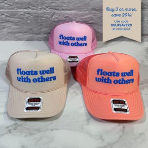 May include: Three trucker hats in light pink, beige, and coral, each featuring a mesh back and the blue text "floats well with others" on the front foam panel. Each hat includes an Otto brand sticker on the brim and a snapback closure.
